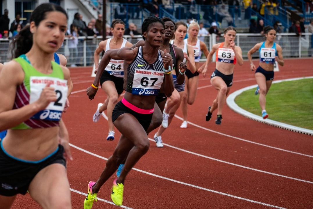 A group of female athletes running on a track during a competitive race, with one athlete in the forefront wearing a black and white striped outfit with the number 67. Other competitors, all focused and determined, are closely following behind. The background shows spectators watching the race from the stands.